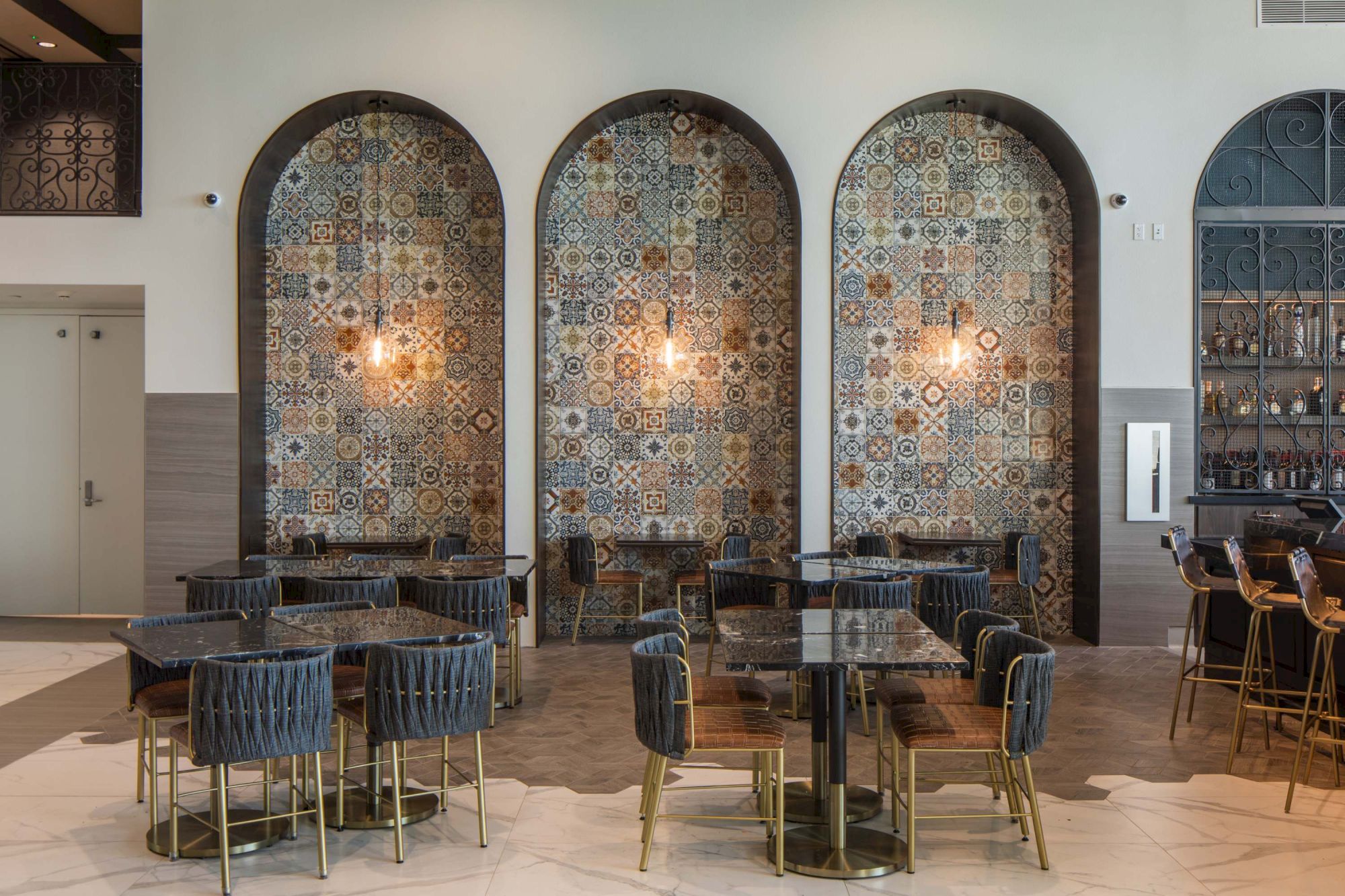 A stylish cafe interior with arched mosaic tile walls, warm lighting, dark upholstered chairs, and round tables with gold legs, creating an elegant dining space.