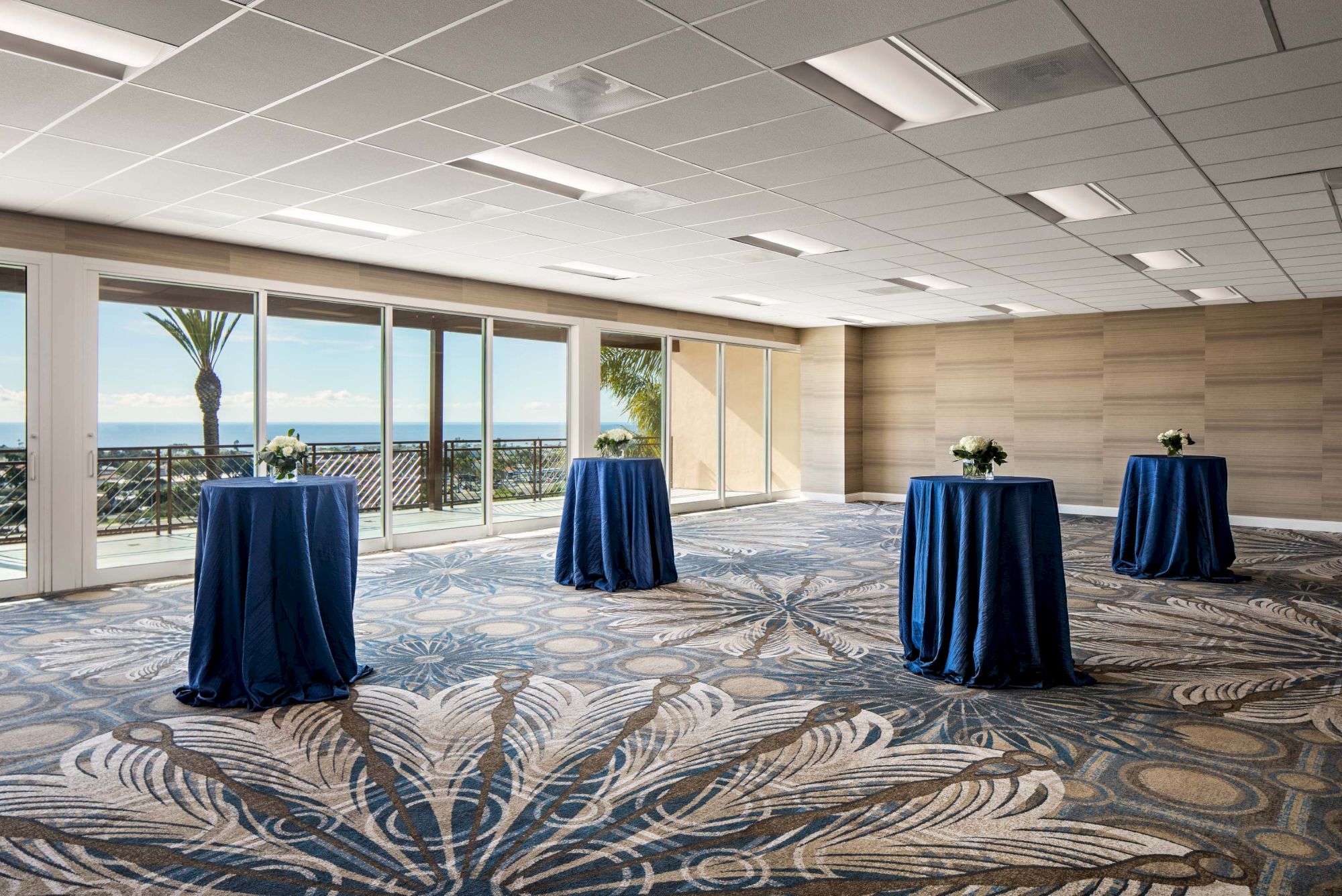 A bright conference room with large windows to a coastal view, and four round high-top tables draped in blue cloth, spaced apart.