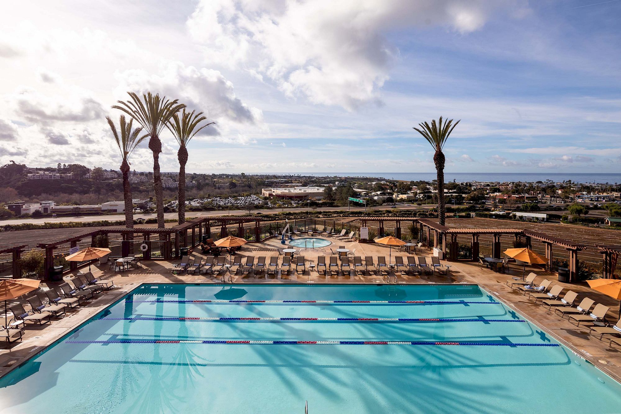 A resort pool area with lounge chairs, two palm trees, and a sunny sky overlooking a distant town, inviting relaxation by the water.