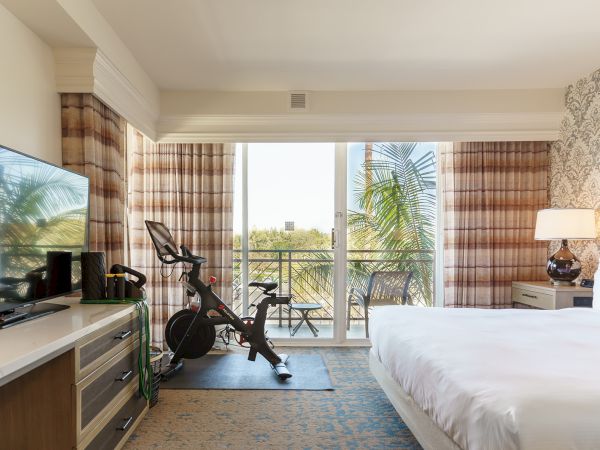 A bright hotel room with a balcony view, featuring a bed, exercise bike, flat-screen TV, dresser, and patterned curtains by large sliding doors to a sunny outdoor patio.