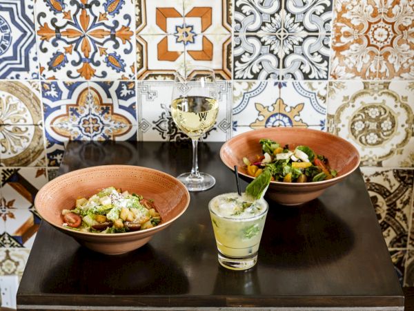 Colorful tiled wall, two bowls with salad, a small drink, and a candle-lit table for two&mdash;cozy cafe vibe.