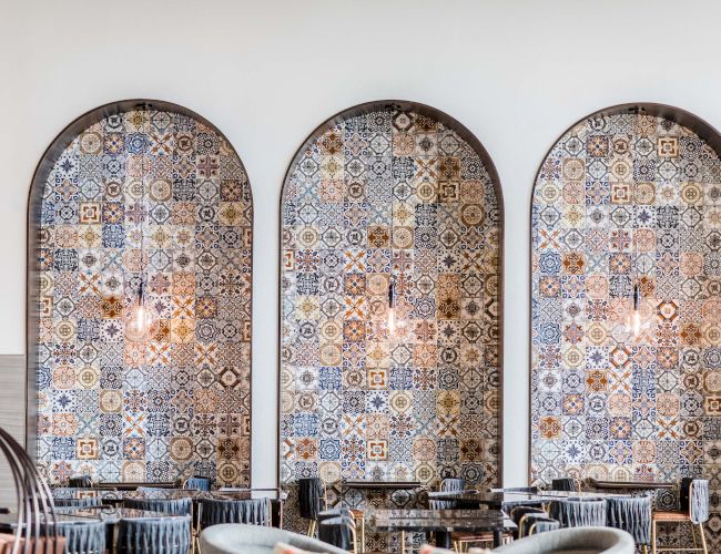 Three arched, patterned tile panels wall-mounted behind a caf&eacute; seating area with chairs and tables in front.