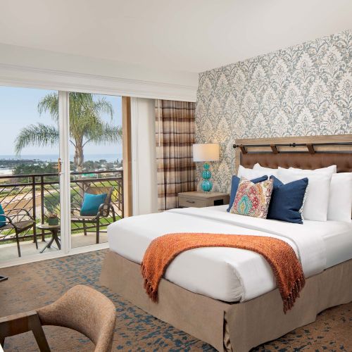 A bright hotel room with a large bed, patterned wallpaper, nightstands, a balcony with seating, and a view of the ocean, all neat and airy.