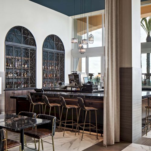 A stylish bar/restaurant interior with a dark wood bar, arched display shelves, and many chairs at tables by large windows, modern lighting, and plants.