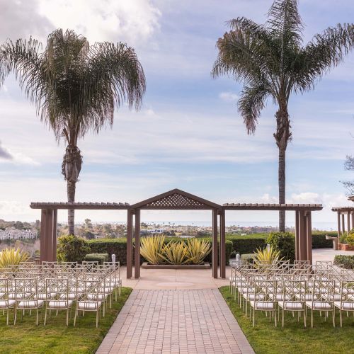 Outdoor wedding setup by a lawn with a wooden arch, two palm trees, white folding chairs arranged in rows, and a distant water view.