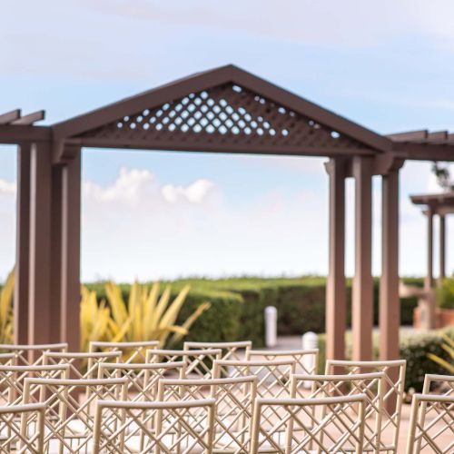 An outdoor wedding setup with wooden arbor, white chairs, and sunny desert landscape in the background, awaiting guests.