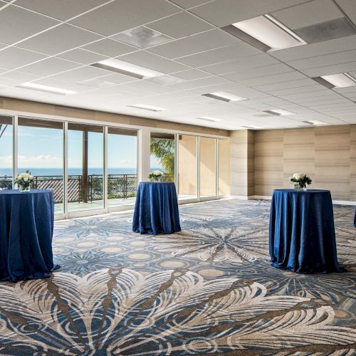 A bright event space with large windows, ocean view, and several tall round tables draped in blue tablecloths, ready for mingling.