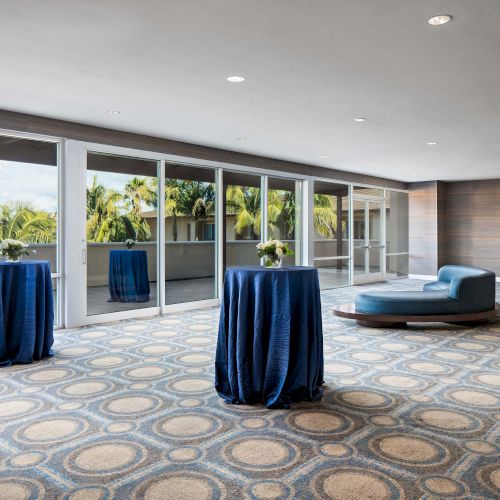 A spacious lounge or event area with patterned carpet, round cocktail tables draped in blue cloth, floor-to-ceiling windows, and a blue curved sofa.