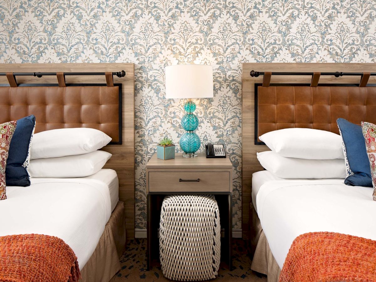 Two twin beds with tufted headboards and a shared nightstand between them; stylish patterned wallpaper, a table lamp, and decorative pillows.