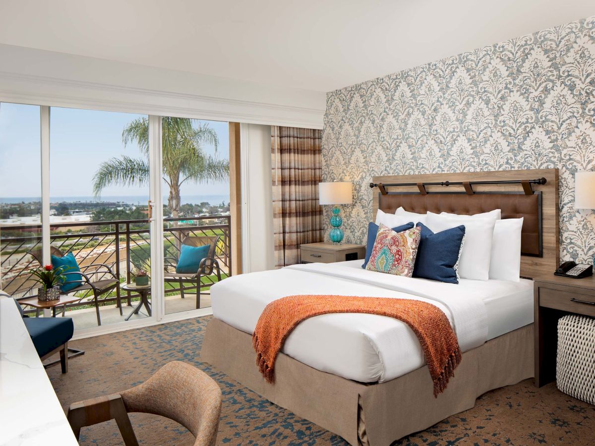 A stylish hotel room with a king bed, patterned wallpaper, teal lamps, and a balcony view of palm trees and an outdoor seating area.