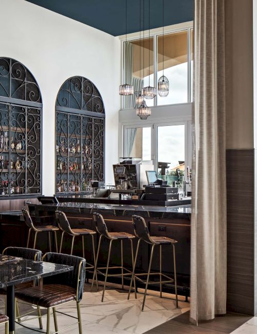 A stylish, modern bar/restaurant interior with a long counter, tall chairs, arched display shelves, and large windows letting in natural light. Top it at 140 characters, always ending the sentence.