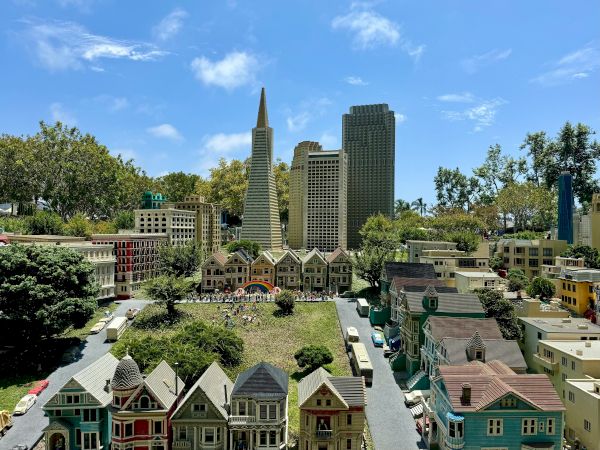 A miniature city with colorful row houses in the foreground, a green park, and tall skyscrapers in the background under a bright blue sky.