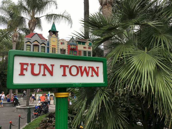 Sign reads &ldquo;FUN TOWN&rdquo; with a small colorful town model on top; tropical plants surround the scene, hints of a carnival or amusement area nearby.