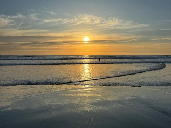 Sunset over calm ocean with a reflective shoreline, warm orange glow, and a lone figure in the distance; serene, tranquil, and picturesque.