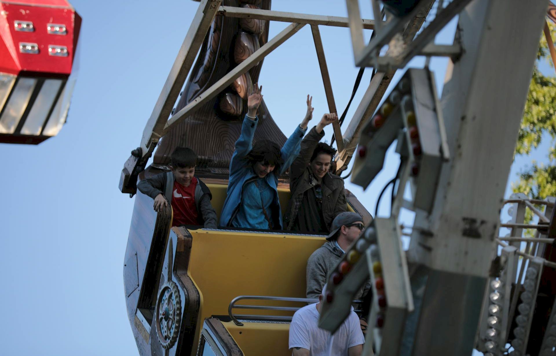 People ride a tilted amusement park ride, looking excited as they lift their arms; a safety harness and support frame are visible.