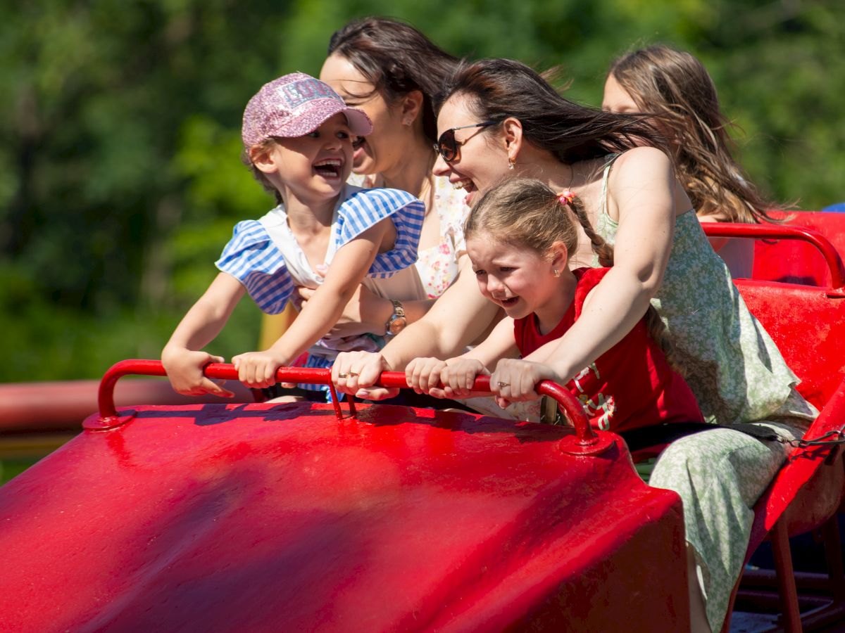 A group of smiling children and an adult ride a red rollercoaster car together, enjoying a sunny amusement park moment.