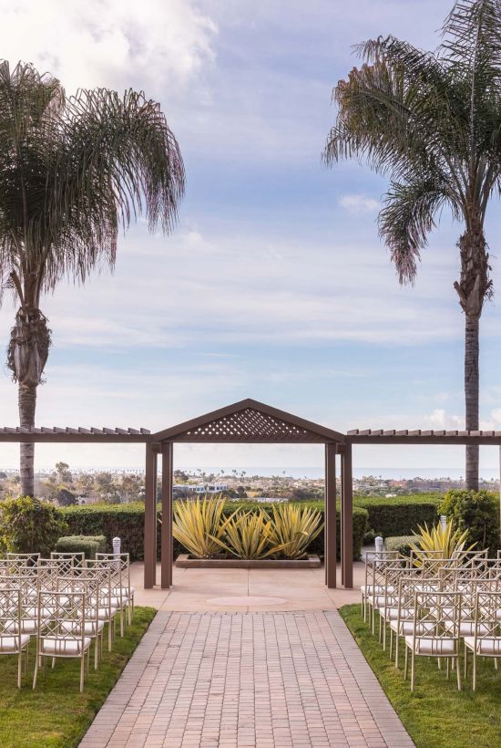 An outdoor wedding setup with rows of white chairs on a grassy lawn, a wooden archway, palm trees, and a lake in the background.