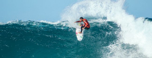 A surfer in a red wetsuit rides a large blue ocean wave, cutting across the crest on a white board, spray flying behind.