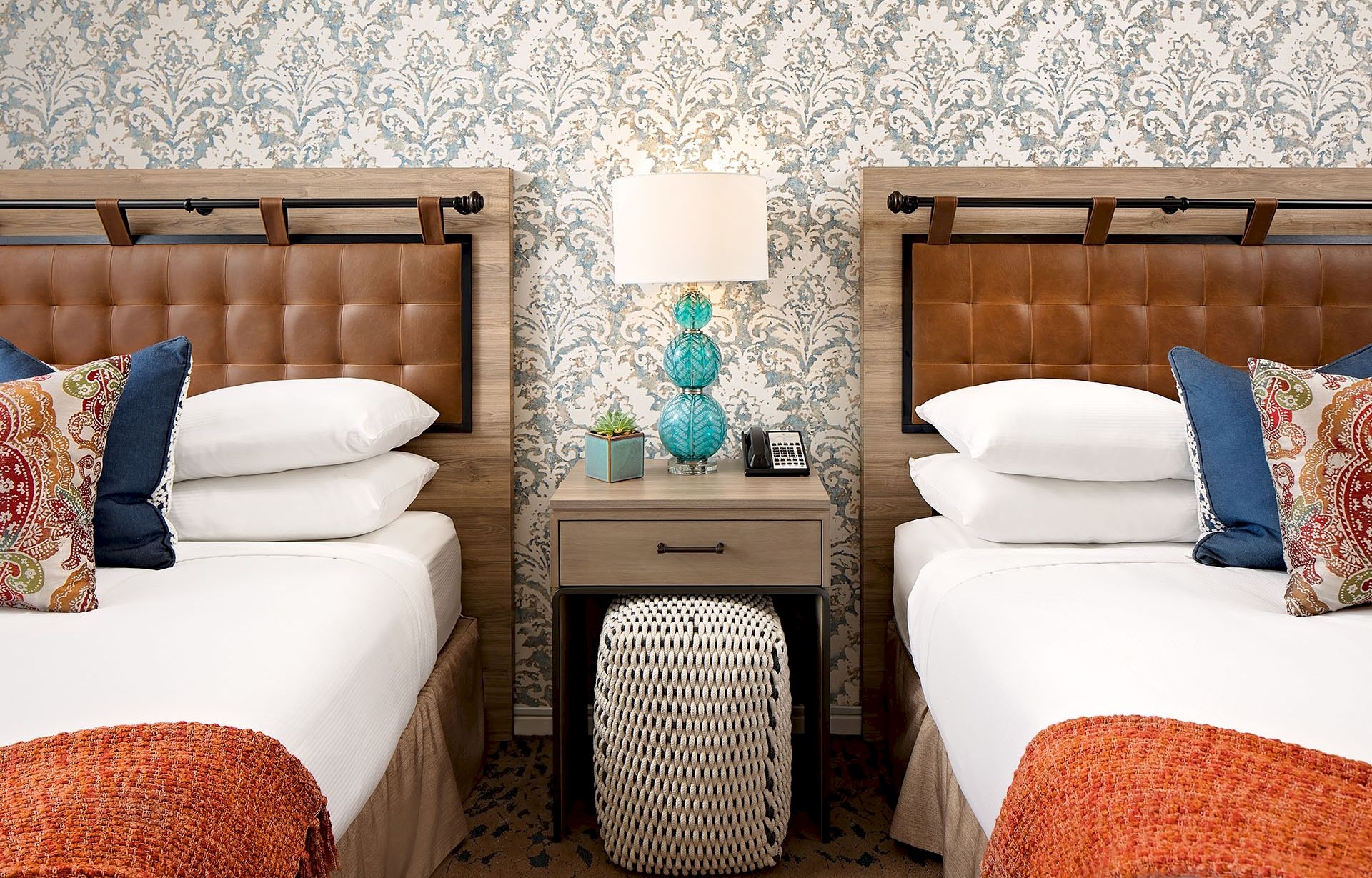 Two twin beds with tufted headboards, a shared nightstand, a turquoise lamp, patterned wallpaper, and decorative pillows in a cozy hotel room.
