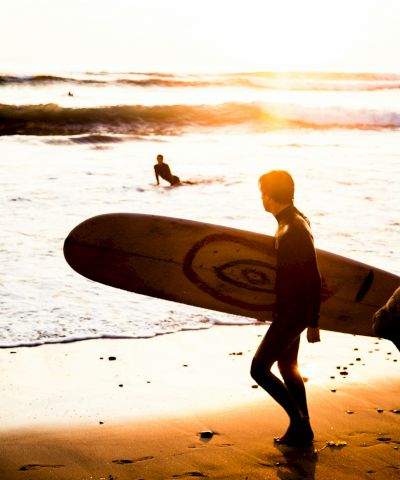 A person carrying a surfboard walks along a sunny beach at sunset, with waves and another surfer in the water, rocks nearby.