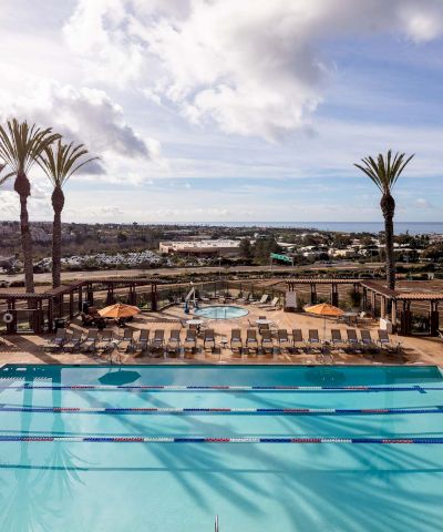 A resort pool with lanes, surrounded by lounge chairs and palm trees, under a bright, partly cloudy sky, overlooking a distant town.