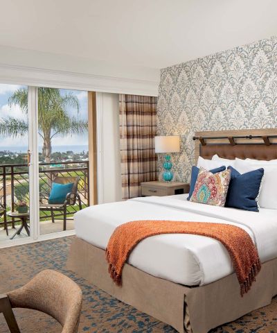 A bright hotel room with a king bed, patterned wallpaper, teal lamps, and a balcony view of palm trees and the ocean, ending with a neat finish.