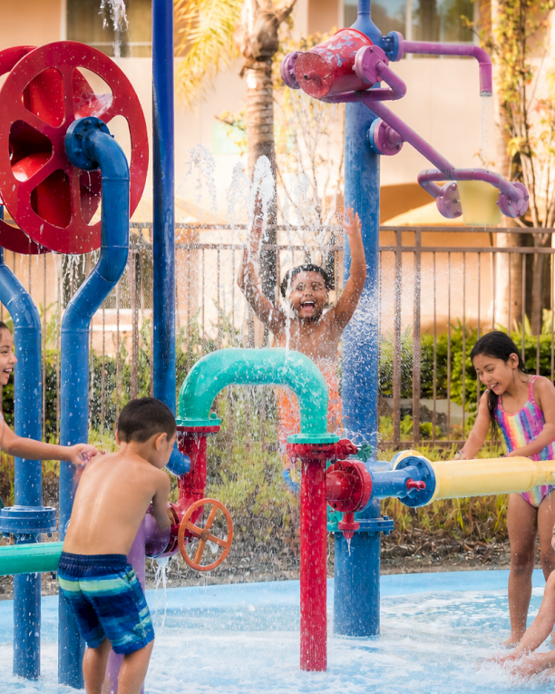 Kids play in a colorful outdoor water park with sprinklers and fountains, splashing in the pool on a sunny day.