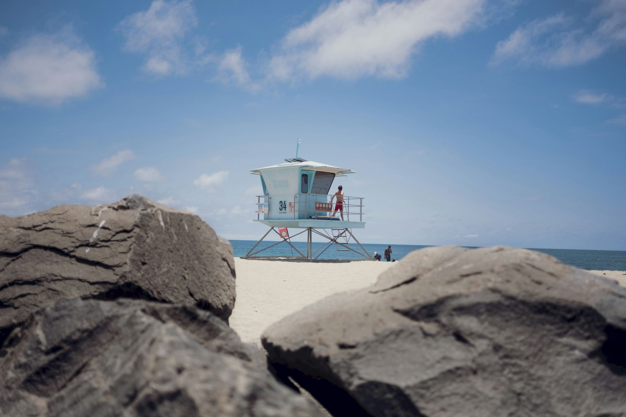 A lifeguard tower stands on a sandy beach with blue sky; large rocks frame the foreground while a few people near the water.